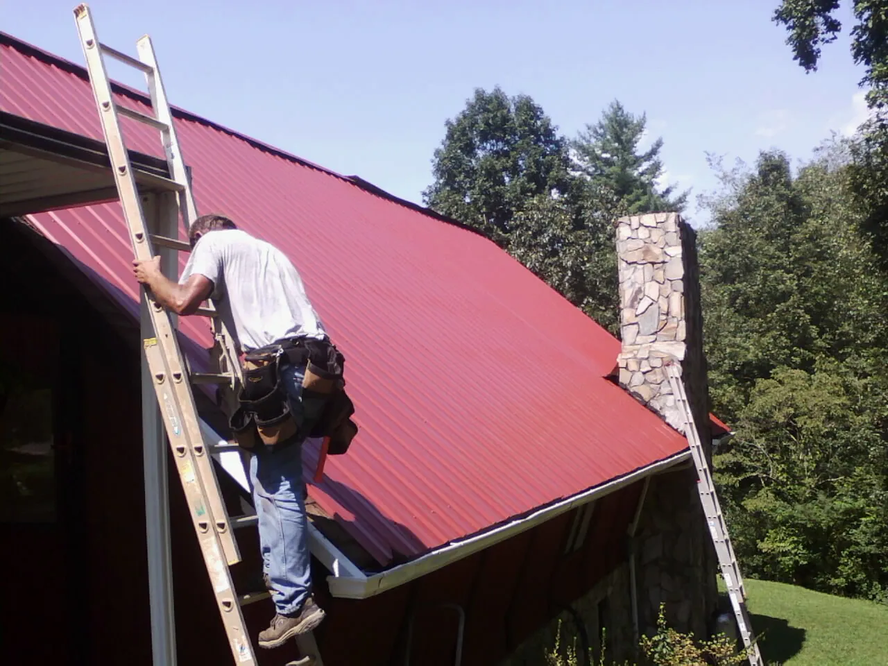 Roofer on a ladder beside a red metal roof and stone chimney — Soffit & Fascia Repair by Roofing Brothers in Clemson